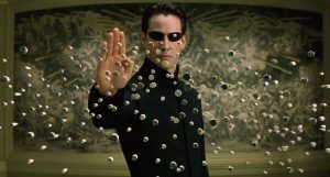 A scene from featuring Neo (Keanu Reeves) in a black trench coat and sunglasses, standing with one hand raised as he stops a barrage of bullets mid-air using his powers. The bullets are suspended around him, emphasizing his control over the Matrix. A detailed mural is visible in the background, adding depth to the scene